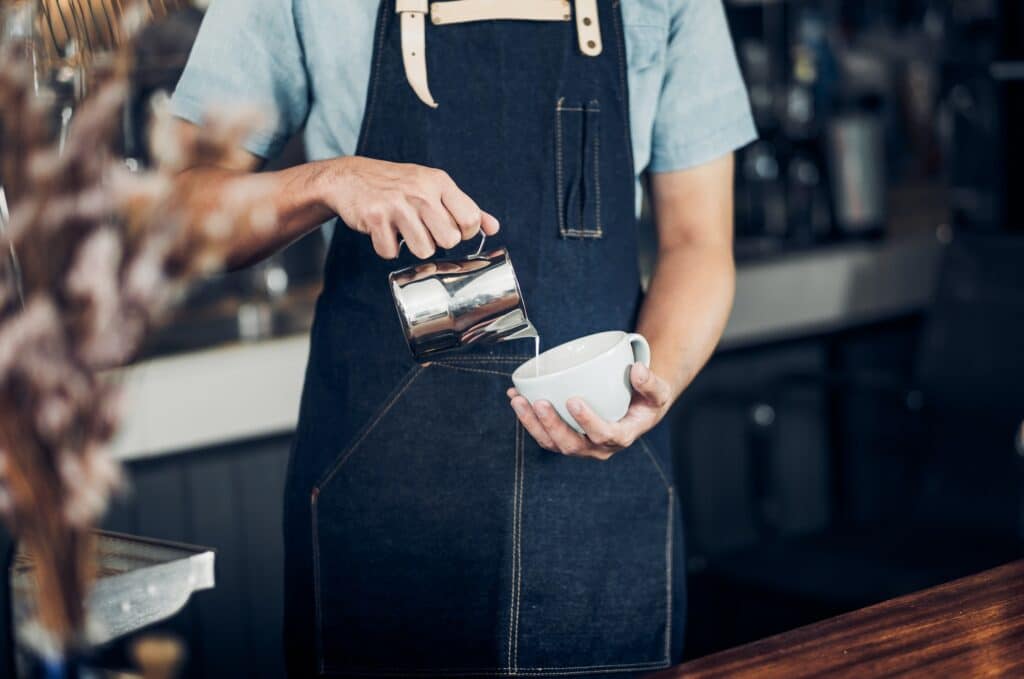 Man barista pour milk into hot coffee cup at counter bar